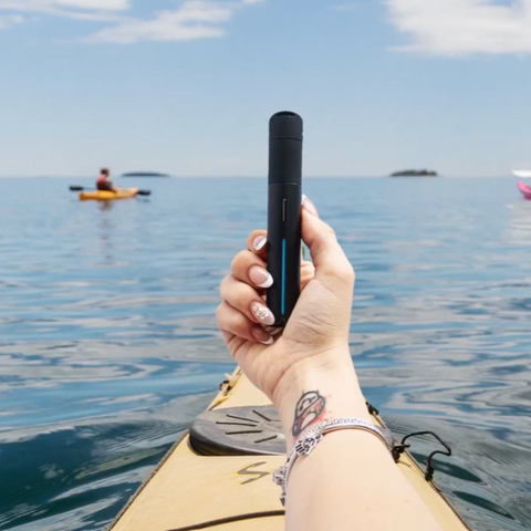 Hand hält den Puffco Pivot Mini Dab Rig Vaporizer auf einem Kajak, umgeben von Wasser und blauen Himmel, während im Hintergrund ein Paddler zu sehe…