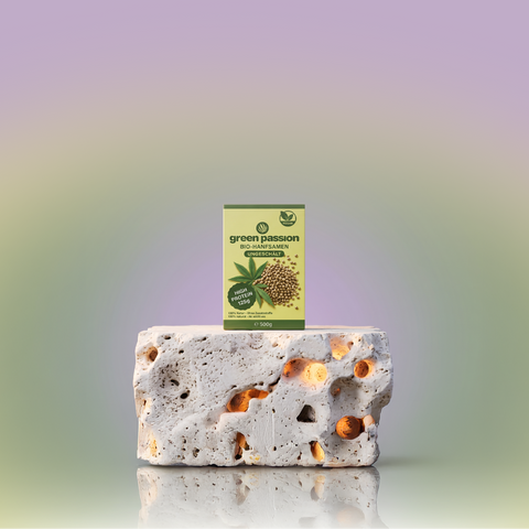A package of Green Passion organic hemp seeds (unpeeled) with a high protein label, displayed on a textured stone surface with a gradient background.