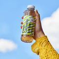 Eine Hand hält eine Flasche Fresh Cannabis Ice Tea von Heimat mit grün-gelbem Etikett vor blauem Himmel.